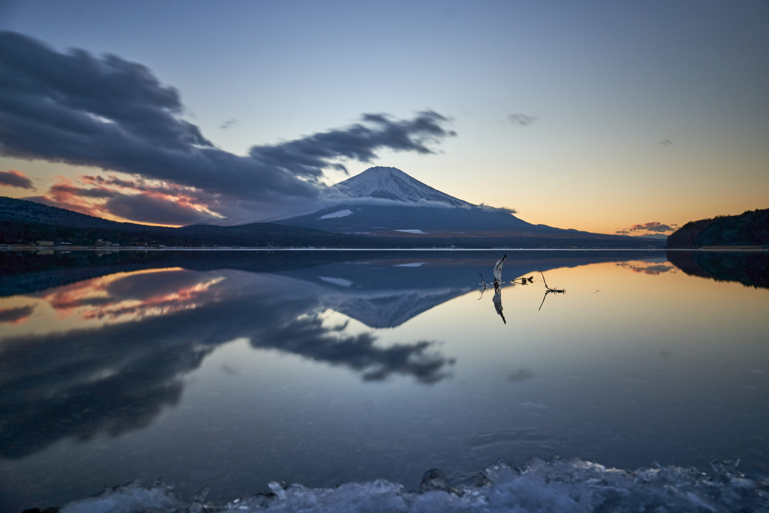 Mount Fuji in the distance, smooth water in the foreground. The mountain is slightly blurry. In the close foreground a stick covered in ice pokes out of the water.