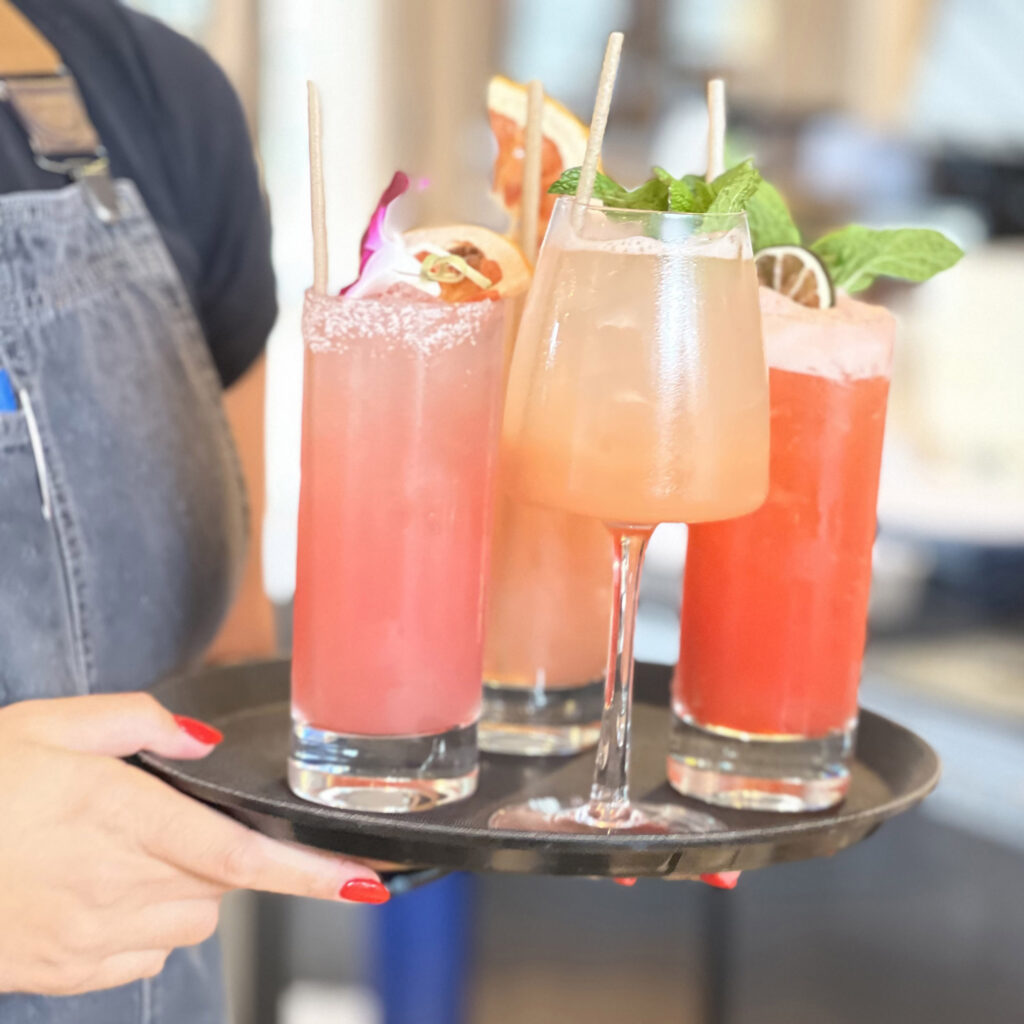 Brightly coiored drinks on a tray held by hands with red nail polish.