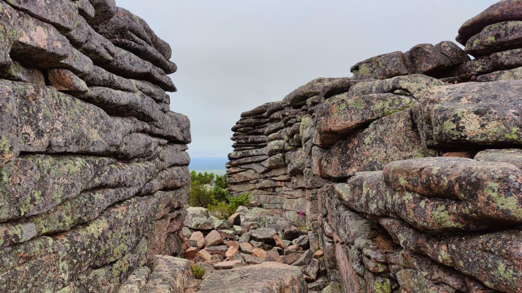 A path through a rock wall.  The wall is made of stratified rock, and the path claims to be made by the wind.