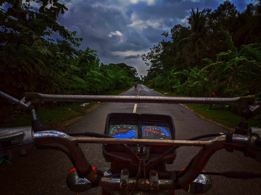 Camera view is sitting on the gas tank of a motorcycle, looking ahead over the handlebars on a straight road, with a cloudy sky after the rain overhead. The wet asphalt glistens, reflecting the dim sunlight, creating a serene and atmospheric scene. The road is narrow with jungle on each side.